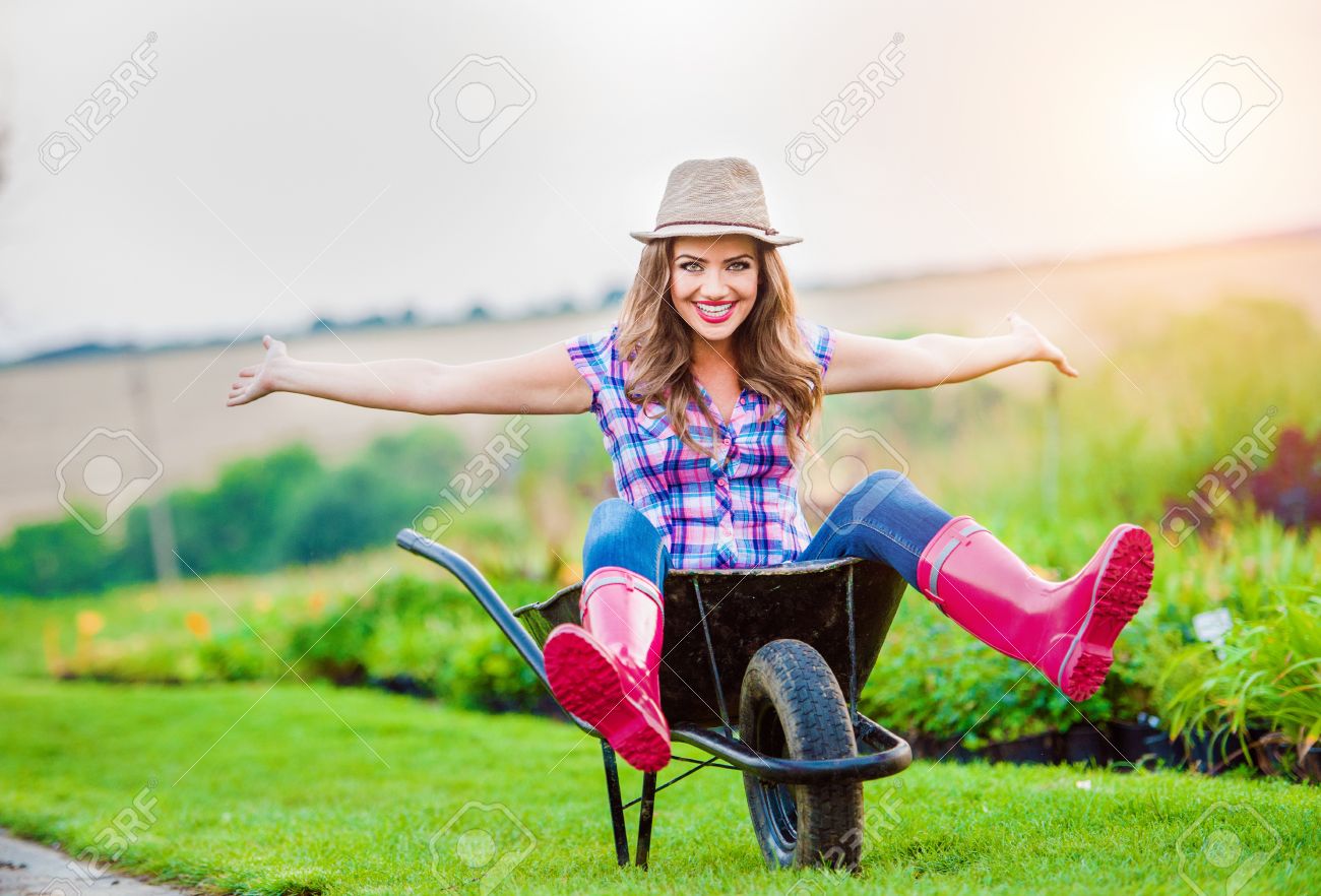 54722716-beautiful-woman-in-rubber-boots-and-hat-sitting-in-wheelbarrow-in-sunny-green-garden.jpg